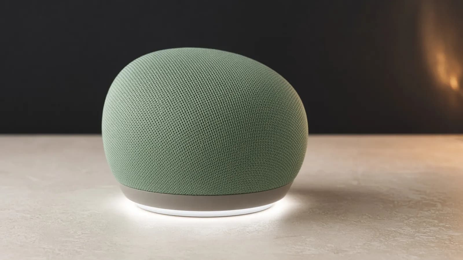 A compact sage green fabric-wrapped spherical smart speaker with a soft white light glowing from its base on a warm stone surface
